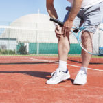 Man on a tennis court reaching for his knee in pain after an injury