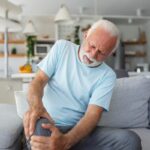 An older man sits on a couch holding his knee in pain.