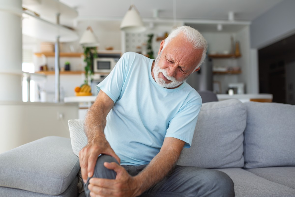 An older man sits on a couch holding his knee in pain.