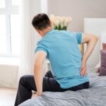 Man sitting on bed holding his lower back in pain, representing chronic back pain that may be treated with endoscopic rhizotomy.