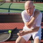 Man sitting on a tennis court holding his shoulder, a common injury that may require rotator cuff surgery.