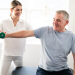 Older man working with a physical therapist using a light weight during shoulder surgery recovery time in a rehab clinic.