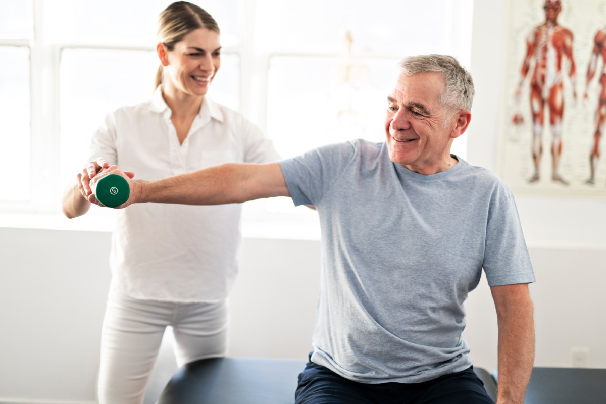 Older man working with a physical therapist using a light weight during shoulder surgery recovery time in a rehab clinic.
