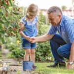 Grandfather crouching in garden with granddaughter picking raspberries after hip replacement recovery, demonstrating restored mobility and active lifestyle.
