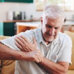 Older man in a t-shirt sitting on a yellow couch, grimacing and experiencing shoulder pain when lifting arm.