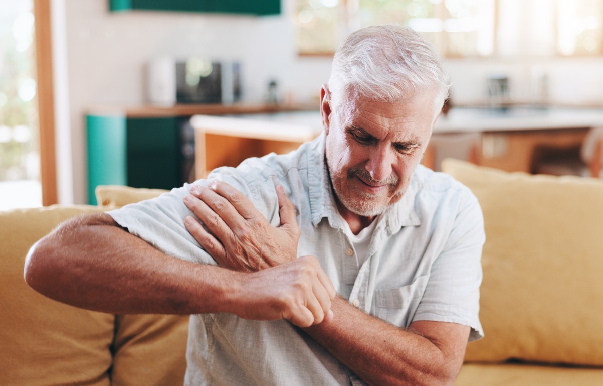 Older man in a t-shirt sitting on a yellow couch, grimacing and experiencing shoulder pain when lifting arm.